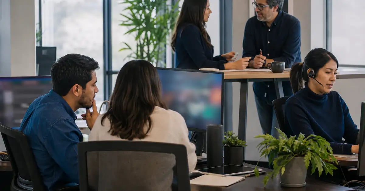 Equipo de agencia latinoamericano trabajando en una oficina abierta: dos personas frente a un monitor compartido, una en llamada y dos conversando de pie
