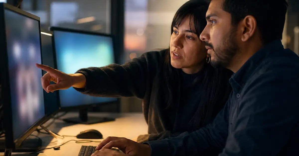 Dos profesionales de TI latinoamericanos trabajando juntos frente a dos monitores durante una tarea técnica de migración de datos