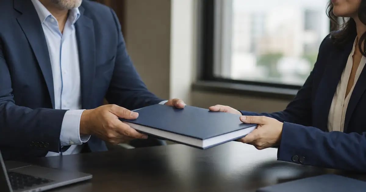 Consultor entregando un documento encuadernado a un cliente al otro lado del escritorio, en el momento del traspaso formal