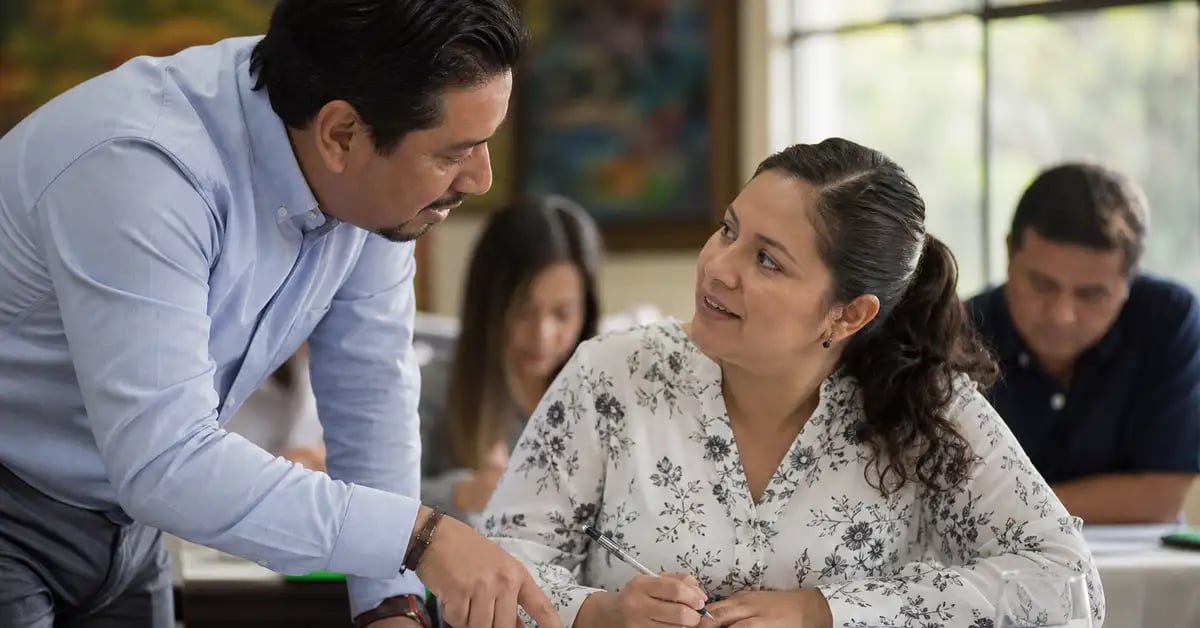 Facilitador latinoamericano agachado junto a una participante, revisando su hoja de trabajo y dándole retroalimentación durante un taller